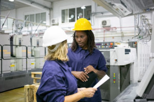 apprenticeship female factory employees in hardhats and overalls standing on plant floor and chatting